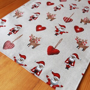 May include: A beige table runner with a repeating pattern of red hearts, reindeer, and Santa Claus figures. The runner is made of a linen-like fabric and has a subtle texture.