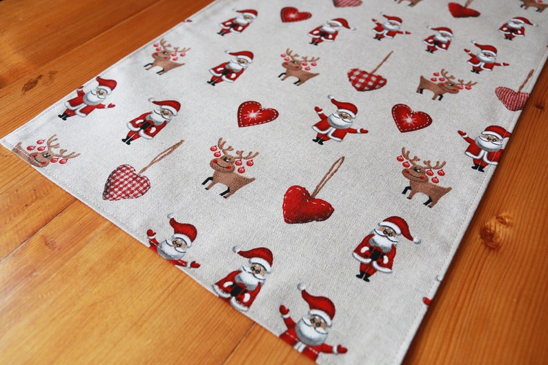 May include: A beige table runner with a repeating pattern of red hearts, reindeer, and Santa Claus figures. The runner is made of a linen-like fabric and has a subtle texture.