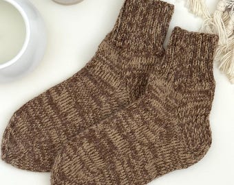 May include: A pair of brown and tan knit socks. The socks are made of a textured yarn and have a ribbed cuff. The socks are lying on a white surface, with a decorative vase and a fringed blanket in the background.