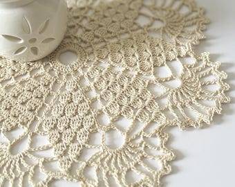 May include: A round, cream-colored crochet doily featuring an intricate, repeating floral-inspired pattern with openwork details. A white ceramic wax warmer with a cutout flower design and a piece of white wax sits on the edge of the doily.