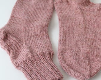 May include: A pair of hand-knitted socks in a dusty rose color. The socks are made of a soft, textured yarn with ribbed cuffs and a detailed heel and toe construction. The socks are laying on a white surface.
