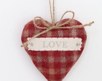 May include: A heart-shaped ornament made of red and tan plaid fabric. A ribbon with the word "LOVE" in silver lettering is attached. The ornament hangs from a twine loop, perfect for Valentine's Day or a romantic gift.