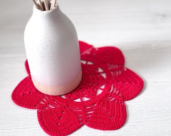May include: A white ceramic vase with reed diffusers sits on a bright red, crocheted doily. The doily has a floral design with a scalloped edge. The vase has a speckled texture and a pink base.