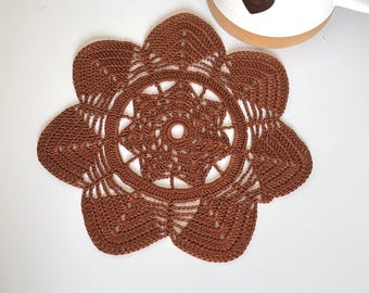 May include: A round, rust-colored crochet doily featuring a six-petaled flower design with intricate stitching and a central circular opening, positioned next to a white ceramic mug decorated with a small, dark brown heart icon.