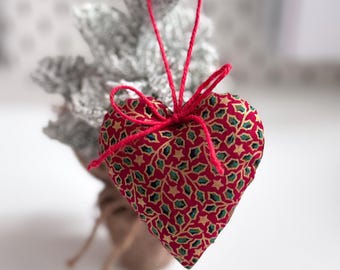 May include: A red heart-shaped Christmas ornament with a gold and green pattern. The ornament is tied with a red string and is hanging from a small, frosted Christmas tree. The ornament is approximately 3 inches wide.