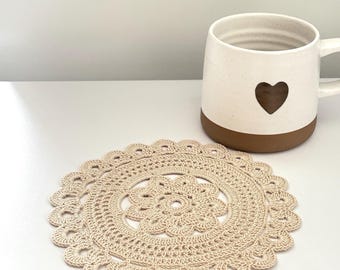 May include: A round, beige crochet doily featuring a central floral pattern surrounded by concentric rings of intricate stitching and a scalloped edge, positioned next to a white ceramic mug with a brown base and a small dark heart icon.