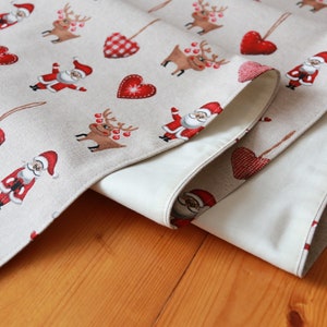 May include: A beige table runner with a Christmas theme featuring red hearts, reindeer, and Santa Claus figures. The runner is folded in half, showing the beige fabric on the underside.