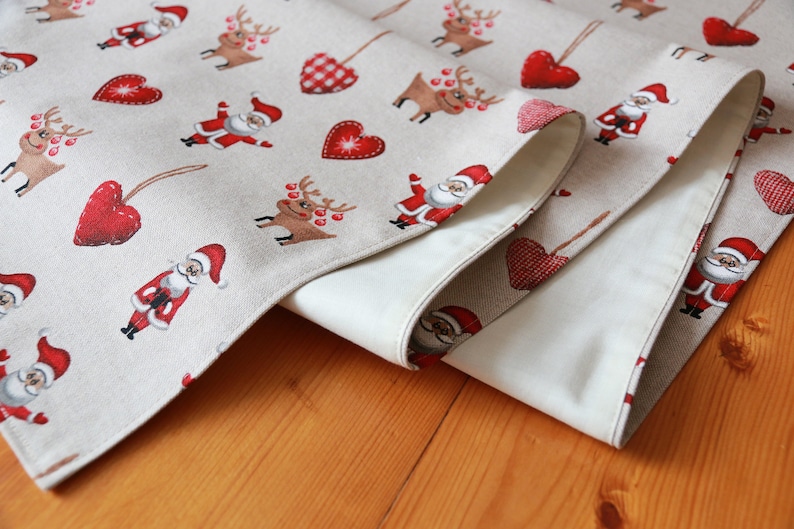 May include: A beige table runner with a Christmas theme featuring red hearts, reindeer, and Santa Claus figures. The runner is folded in half, showing the beige fabric on the underside.