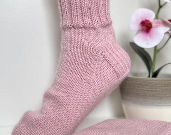 May include: A pair of light pink hand-knitted socks featuring a ribbed cuff and a smooth stockinette stitch pattern. One sock is displayed upright on a foot form, while the other is folded neatly beside it, showcasing the soft yarn texture.