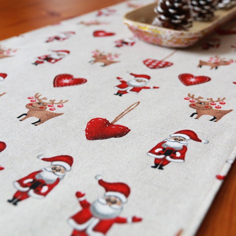 May include: A beige linen table runner with a repeating pattern of red hearts, reindeer, and Santa Claus figures. The runner is decorated with a festive Christmas theme.