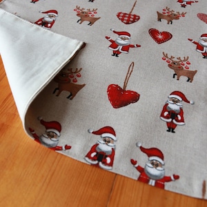 May include: A beige table runner with a Christmas theme. The runner features a repeating pattern of red hearts, reindeer, and Santa Claus figures. The runner is made of a linen-like fabric.
