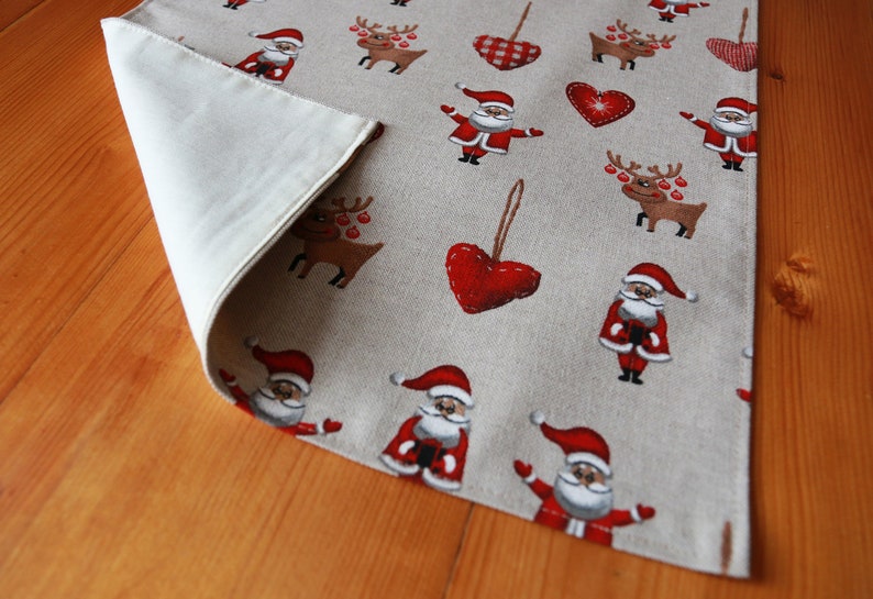 May include: A beige table runner with a Christmas theme. The runner features a repeating pattern of red hearts, reindeer, and Santa Claus figures. The runner is made of a linen-like fabric.