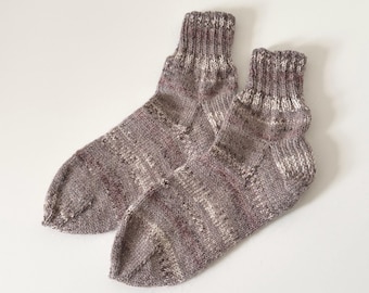 May include: A pair of hand-knitted socks in shades of gray, mauve, and white. The socks have ribbed cuffs and a patterned design throughout the body. The socks are made of a soft, textured yarn, perfect for cozy wear.