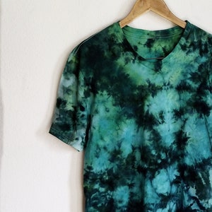 K&ouml;nnte beinhalten: Ein kurz&auml;rmeliges T-Shirt mit gr&uuml;nem und t&uuml;rkisfarbenem Batikmuster h&auml;ngt an einem Holzb&uuml;gel. Das Shirt hat einen runden Ausschnitt und ein einzigartiges, wirbelndes Muster. &Uuml;ber dem Shirt befindet sich ein Holzschild mit der Aufschrift "Tye Me Up Designs Est. 2017."