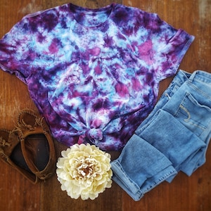 May include: A tie-dye t-shirt in shades of purple, blue, and white, with a pair of blue jeans, brown leather moccasins, and a white flower.