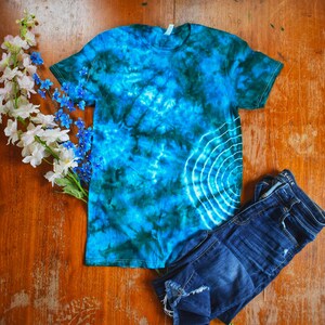 May include: A tie-dye t-shirt in shades of blue and green, with a white circular design, is displayed with a pair of blue denim shorts and white and blue flowers on a wooden surface. The t-shirt is short-sleeved.