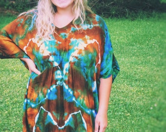 May include: A tie-dye maxi dress in shades of brown, blue, green, and white. The dress has a V-neck and loose sleeves. A brown hat with a dark blue band is worn. The background is a green lawn and trees.