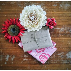 Op de afbeelding: Een stilleven met een in bruin papier verpakt pakket, vastgebonden met touw, op een roze bloemenstof. Omringd door een rode zonnebloem, een cr&egrave;mekleurige pioenroos en een kleine rode roos, alles op een rustiek houten oppervlak.