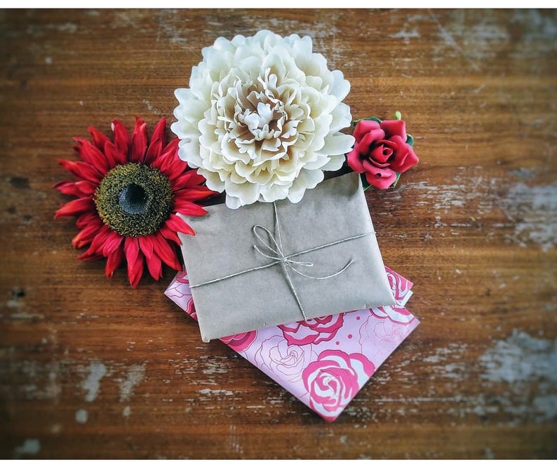 Op de afbeelding: Een stilleven met een in bruin papier verpakt pakket, vastgebonden met touw, op een roze bloemenstof. Omringd door een rode zonnebloem, een cr&egrave;mekleurige pioenroos en een kleine rode roos, alles op een rustiek houten oppervlak.
