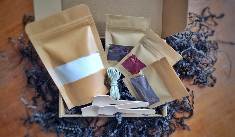 May include: A craft kit in a cardboard box, containing several kraft paper pouches with colored contents, wooden spoons, and a bundle of string. The pouches have clear windows showing the colors inside. The box is filled with black paper shreds.