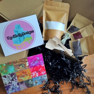 May include: An open cardboard box filled with craft supplies. Inside are small brown paper bags, wooden spoons, string, and a card with color swatches labeled "Bronze," "Marigold," and "Blue Gray." A card with the logo "EyeMeUpDesigns" is also visible.