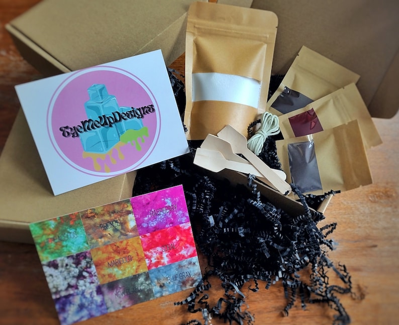 May include: An open cardboard box filled with craft supplies. Inside are small brown paper bags, wooden spoons, string, and a card with color swatches labeled "Bronze," "Marigold," and "Blue Gray." A card with the logo "EyeMeUpDesigns" is also visible.