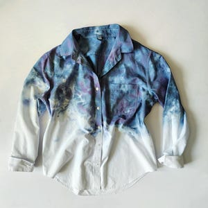 May include: A button-down shirt with long sleeves, featuring a tie-dye design. The top portion of the shirt is a blend of blue and purple hues, transitioning to white at the bottom. The shirt has a single chest pocket and a classic collar.