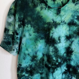 K&ouml;nnte beinhalten: Ein gr&uuml;n-blaues T-Shirt mit Batikmuster und V-Ausschnitt. Das Shirt h&auml;ngt an einem Holzkleiderb&uuml;gel vor einer wei&szlig;en Wand. Das Batikmuster weist Gr&uuml;nt&ouml;ne, T&uuml;rkis und Schwarz auf und erzeugt ein einzigartiges, abstraktes Design. Die kurzen &Auml;rmel des Shirts sind sichtbar.