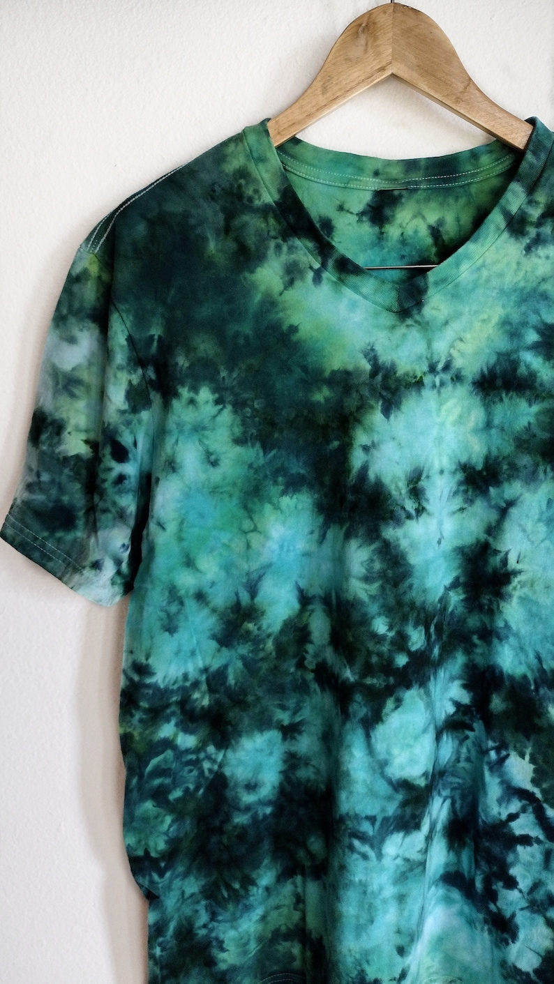 K&ouml;nnte beinhalten: Ein gr&uuml;n-blaues T-Shirt mit Batikmuster und V-Ausschnitt. Das Shirt h&auml;ngt an einem Holzkleiderb&uuml;gel vor einer wei&szlig;en Wand. Das Batikmuster weist Gr&uuml;nt&ouml;ne, T&uuml;rkis und Schwarz auf und erzeugt ein einzigartiges, abstraktes Design. Die kurzen &Auml;rmel des Shirts sind sichtbar.