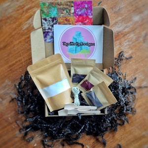May include: An open cardboard box filled with craft supplies. The box contains small brown paper bags, wooden spoons, a bundle of string, and a card with the text "EyeMeUpDesigns". The box is surrounded by black paper shreds.