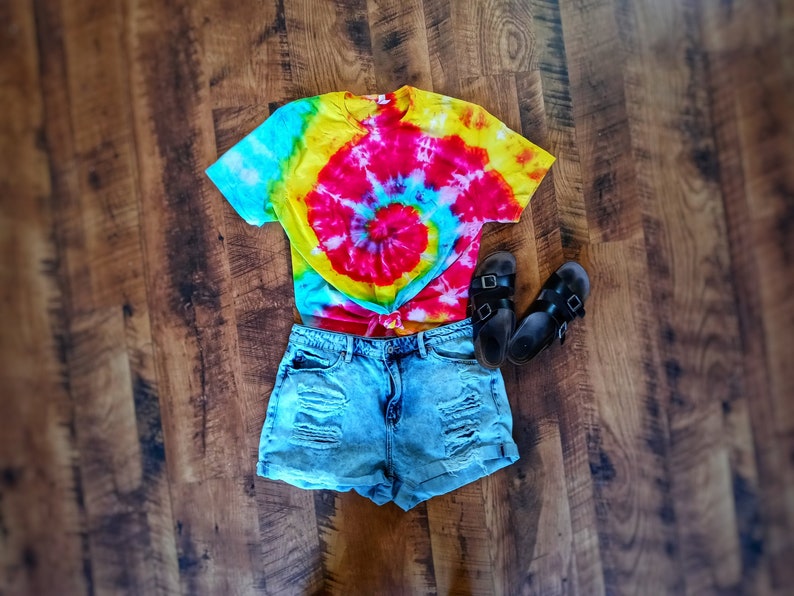 Op de afbeelding: Een kleurrijk tie-dye t-shirt met een spiraalontwerp in rood, geel en blauw, gecombineerd met distressed denim shorts en zwarte sandalen. De outfit is geplaatst op een houten oppervlak, wat een casual, zomerse stijl suggereert.