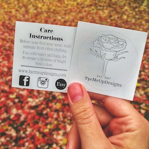 Puede incluir: Una mano sosteniendo dos tarjetas blancas con instrucciones de cuidado y un logotipo de rosa. Las tarjetas incluyen la direcci&oacute;n del sitio web, iconos de redes sociales y el texto "TyeMeUpDesigns". El fondo es una vista borrosa de hojas de oto&ntilde;o.