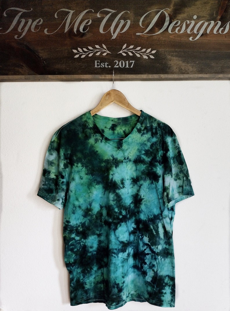 K&ouml;nnte beinhalten: Ein kurz&auml;rmeliges T-Shirt mit gr&uuml;nem und t&uuml;rkisfarbenem Batikmuster h&auml;ngt an einem Holzb&uuml;gel. Das Shirt hat einen runden Ausschnitt und ein einzigartiges, wirbelndes Muster. &Uuml;ber dem Shirt befindet sich ein Holzschild mit der Aufschrift "Tye Me Up Designs Est. 2017."