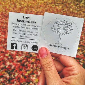 Op de afbeelding: Een hand houdt twee witte kaarten vast met verzorgingsinstructies en een rozenlogo. De kaarten bevatten het websiteadres, social media-iconen en de tekst "TyeMeUpDesigns". De achtergrond is een wazig beeld van herfstbladeren.