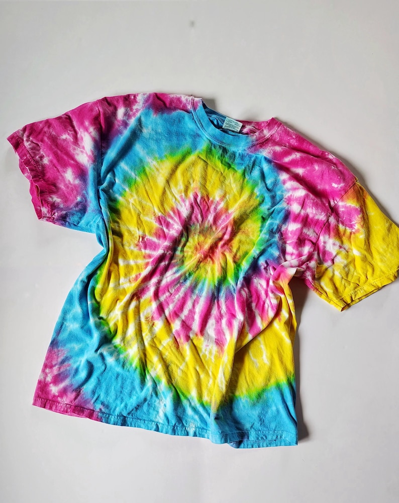 Op de afbeelding: Een kleurrijk tie-dye T-shirt met een spiraalpatroon. Het shirt heeft een mix van levendige kleuren, waaronder roze, geel, blauw en groen. Het shirt met korte mouwen ligt plat op een witte ondergrond.