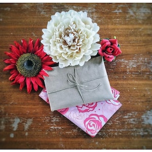 Puede incluir: Naturaleza muerta con un paquete envuelto en papel marr&oacute;n y atado con hilo, sobre una tela rosa con estampado floral. Rodeado por un girasol rojo, una peon&iacute;a crema y una peque&ntilde;a rosa roja, todo sobre una superficie de madera r&uacute;stica.