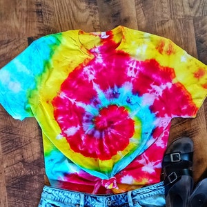 Op de afbeelding: Een levendig tie-dye T-shirt met een spiraalpatroon, met een mix van heldere kleuren zoals rood, geel en blauw. Het shirt is in de taille geknoopt en gecombineerd met een spijkerbroekshort en zwarte sandalen.