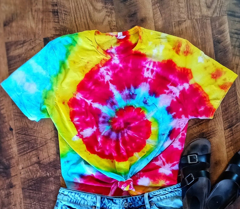 Op de afbeelding: Een levendig tie-dye T-shirt met een spiraalpatroon, met een mix van heldere kleuren zoals rood, geel en blauw. Het shirt is in de taille geknoopt en gecombineerd met een spijkerbroekshort en zwarte sandalen.