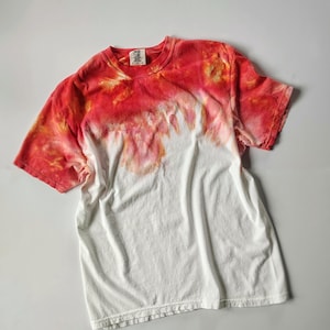 Puede incluir: Camiseta blanca con dise&ntilde;o tie-dye. La parte superior est&aacute; te&ntilde;ida en tonos rojos y naranjas, difumin&aacute;ndose en una secci&oacute;n inferior blanca. Cuenta con mangas cortas y cuello redondo.