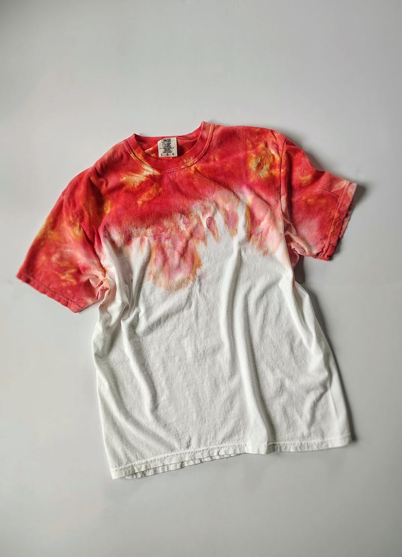 Puede incluir: Camiseta blanca con dise&ntilde;o tie-dye. La parte superior est&aacute; te&ntilde;ida en tonos rojos y naranjas, difumin&aacute;ndose en una secci&oacute;n inferior blanca. Cuenta con mangas cortas y cuello redondo.