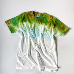 Puede incluir: Una camiseta blanca con un dise&ntilde;o tie-dye. La parte superior de la camiseta est&aacute; te&ntilde;ida en tonos verdes y marrones, pasando al azul y luego al blanco. La camiseta est&aacute; hecha de un material suave y c&oacute;modo.
