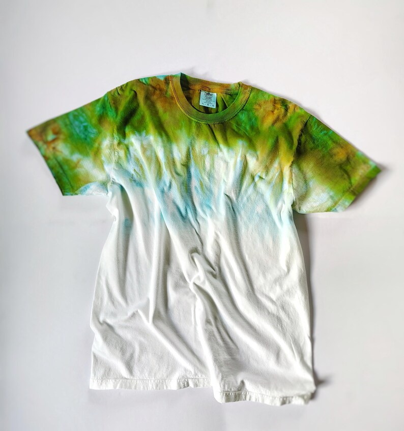 Puede incluir: Una camiseta blanca con un dise&ntilde;o tie-dye. La parte superior de la camiseta est&aacute; te&ntilde;ida en tonos verdes y marrones, pasando al azul y luego al blanco. La camiseta est&aacute; hecha de un material suave y c&oacute;modo.
