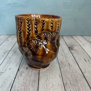 May include: A handcrafted ceramic mug with a brown glaze. The mug features a textured surface with embossed floral and leaf patterns. The mug is approximately 4 inches tall and 4 inches wide.