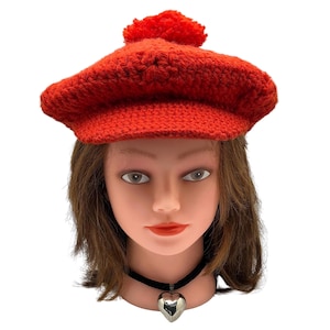May include: A bright red, crocheted beret with a pom-pom on top. The hat is displayed on a mannequin head with brown hair, a black choker, and a silver heart pendant. The hat's texture is visible, showcasing the handmade quality.