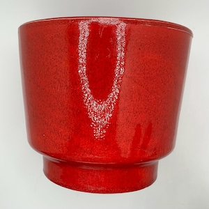 May include: A bright red ceramic plant pot with a textured finish. The pot has a cylindrical shape with a flared rim and a pedestal base. The glossy surface enhances the rich colour, making it a striking decorative piece for plants or home decor.