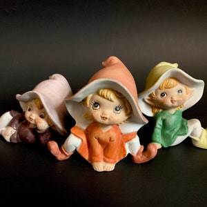 May include: Three ceramic figurines of elves wearing pointed hats. The elves are wearing different coloured outfits: pink, orange, and green. They are all sitting with their legs crossed and have painted facial features.