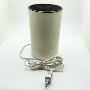May include: A cream-colored, cylindrical appliance with a black interior rim. A white power cord with a two-prong plug is attached. The cord is secured with a brown rubber band. The appliance is likely a beauty product warmer.