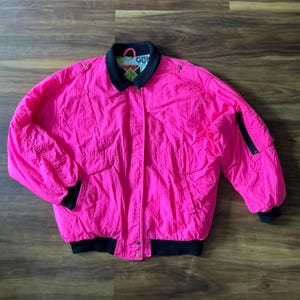 Könnte beinhalten: Leuchtend pinke Vintage-Jacke mit schwarzem Kragen, Bündchen und Saum. Die Jacke hat zwei Vordertaschen, einen Reißverschluss am linken Ärmel und Druckknöpfe. Auf dem Kragen befindet sich ein kleines Logo. Eine Jacke im Retro-Stil.