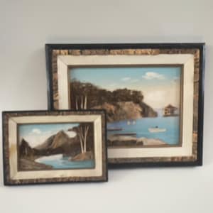 May include: Two framed landscape paintings. The larger painting depicts a coastal scene with a blue sky, water, and a small boat. The smaller painting shows a mountain and river scene. Both paintings have decorative frames.