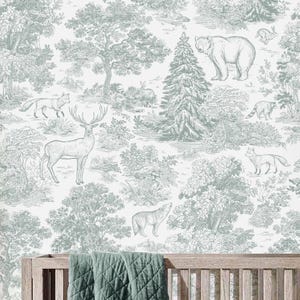 May include: A nursery interior featuring a wooden cot, chest of drawers, and a white armchair. The cot has a green quilted blanket. The wallpaper depicts a forest scene with deer, bears, and trees. A soft toy and cushion are on the chair.